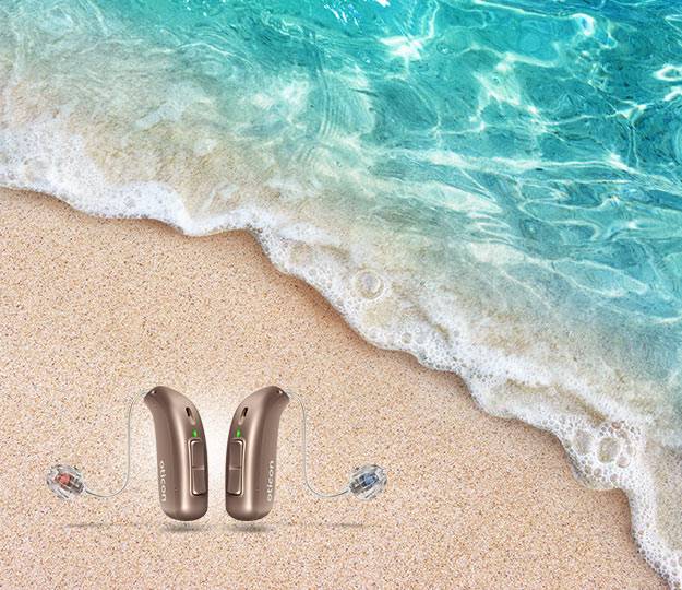 Two Oticon hearing aids rest on sandy beach near turquoise ocean waves, emphasizing hearing care in serene outdoor settings. Oticon branding is visible on each device.