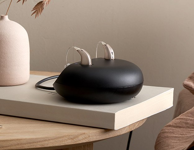 Rechargeable hearing aids resting on a sleek black charging case placed on a book on a wooden table.
