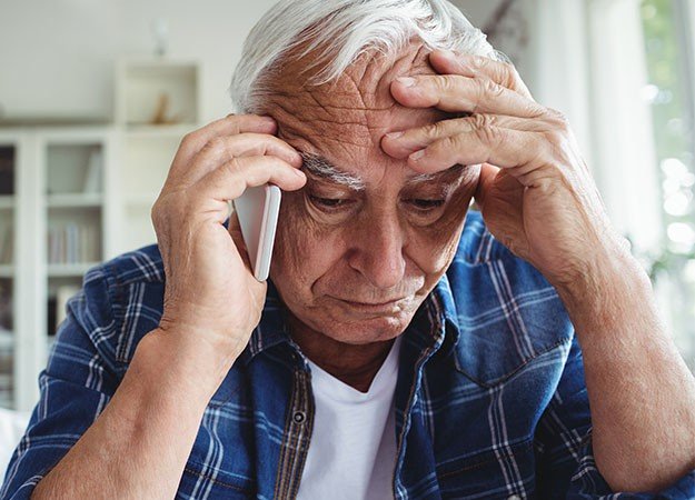 Homem idoso segurando a cabeça com expressão de desconforto enquanto fala ao telefone.