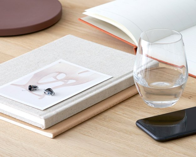 Due apparecchi acustici discreti sono posati su un libro chiaro, accanto a un bicchiere d’acqua, uno smartphone e un’agenda aperta, su una scrivania in legno.