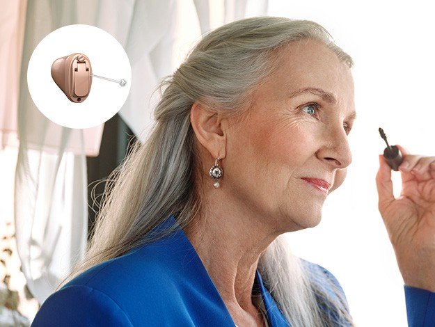 Aparelho auditivo discreto sendo mostrado por uma mulher de cabelos grisalhos em ambiente iluminado por luz natural. No canto superior esquerdo, destaque para o design interno do dispositivo.