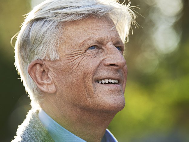 Carl, en ældre mand med gråt hår, står udendørs i sollys med fokus på hans øre og omgivet af grønne nuancer.