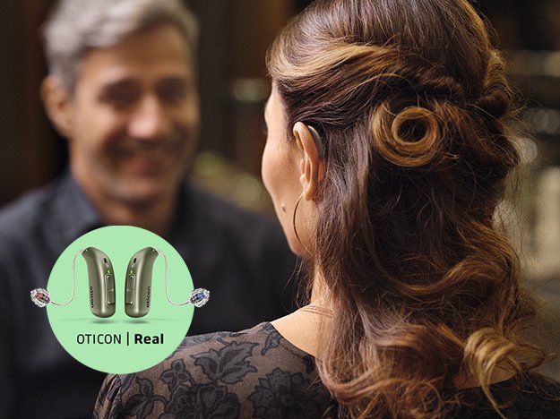 Een vrouw met opgestoken haar draagt een onzichtbare Oticon Real hoorapparaat tijdens een gesprek in een stijlvolle binnenruimte. Een inset toont twee Oticon Real hoortoestellen met tekst: 'OTICON | Real'.