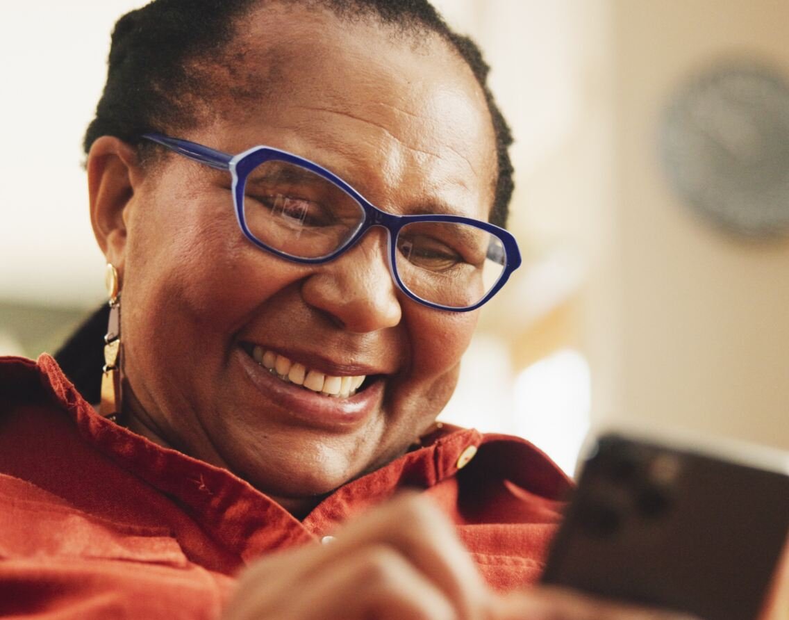 A person interacts with a smartphone while wearing a discreet, blue hearing aid. The setting appears indoors, with soft lighting and a blurred wall clock in the background.