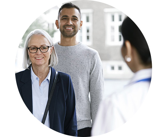An older woman and a younger man stand in front of a healthcare professional in a white coat, possibly an audiologist, outside a building resembling a hearing clinic. No visible text.