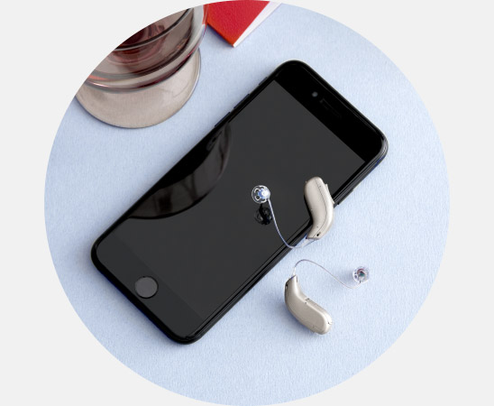 Zwei beige Hörgeräte liegen neben einem schwarzen Smartphone auf einer hellblauen Oberfläche; im Hintergrund ein Glas und ein rotes Buch. Keine Marke erkennbar, typische Hörklinikumgebung.