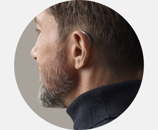 Ein Hinter-dem-Ohr-Hörgerät ist am rechten Ohr eines Mannes befestigt, der einen dunkelblauen Pullover trägt. Der Hintergrund ist neutral und hebt das Hörgerät hervor.