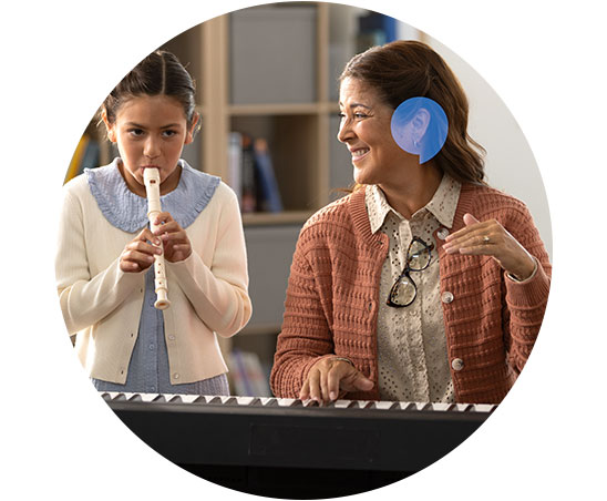 Una donna indossa un apparecchio acustico azzurro mentre suona una tastiera elettronica accanto a una bambina che suona un flauto dolce in un ambiente scolastico.