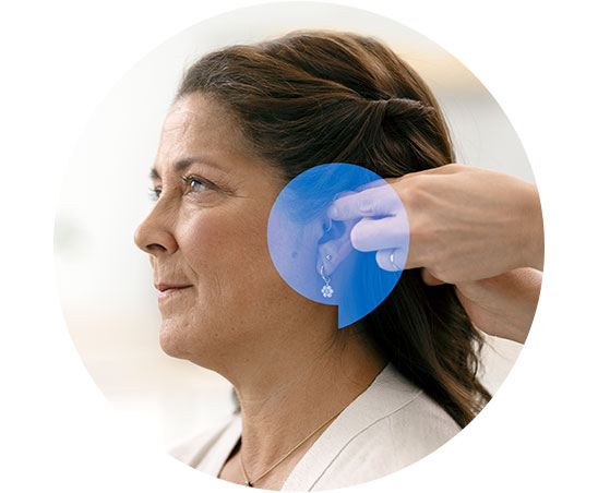 Een audioloog plaatst een onzichtbaar gehoorapparaat achter het oor van een vrouw met bruin haar in een lichte, klinische omgeving.