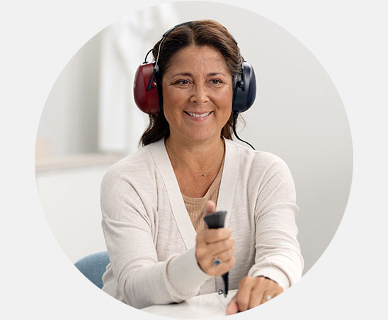 Una donna indossa grandi cuffie rosse e blu, tiene un pulsante manuale e sembra eseguire un test dell’udito in un centro acustico Audika, in un ambiente luminoso.