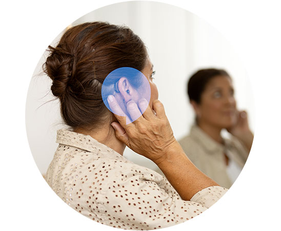 Een vrouw past een hoorapparaat aan haar oor aan, met een close-up van het apparaat. Op de achtergrond zit een persoon in een moderne hoorcentrumomgeving, gezicht onscherp gemaakt.