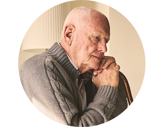 Ein älterer Mann trägt einen grauen Strickpullover, stützt sich auf einen Holzstock und sitzt in einem warm beleuchteten Innenraum mit einer weissen Lampe im Hintergrund.