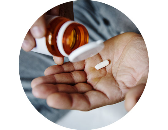 A hand holds a white pill while another hand pours more from an orange prescription bottle, set against an indoor background. 