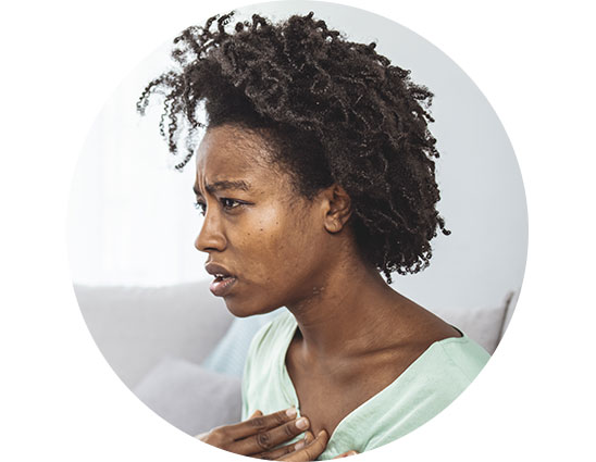 A person with natural, curly hair is seated indoors, wearing a light green top, with a hand placed on their chest. The background shows a softly lit couch and cushions.