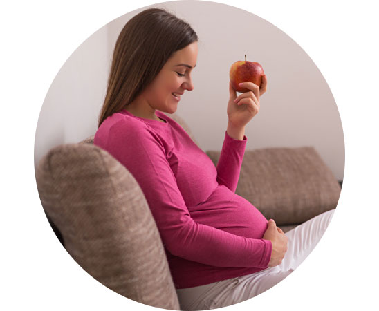 Zwangere vrouw in een roze trui zit op de bank en eet een rode appel.