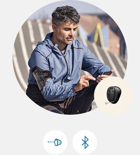 A man in a blue jacket uses a smartphone outdoors on a sunny day, with an Oticon hearing aid icon displayed nearby. Bluetooth and in-ear hearing aid symbols appear below.