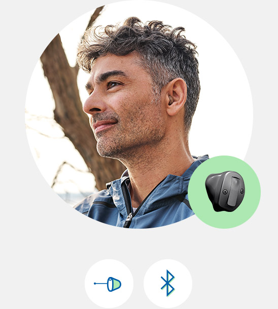 En mand med kort, krøllet hår iført en blå jakke ses udendørs med træer i baggrunden. Ikoner viser Bluetooth-forbindelse og et lille, diskret høreapparat fra Audika.