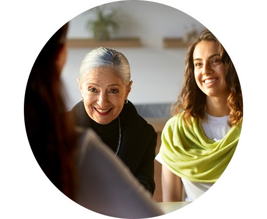 To ældre kvinder sidder ved et bord i en samtale med en person vendt mod kameraet. Baggrunden viser slørede hylder og planter i en hyggelig klinikindretning. Ingen synlige høreapparater eller tekst.