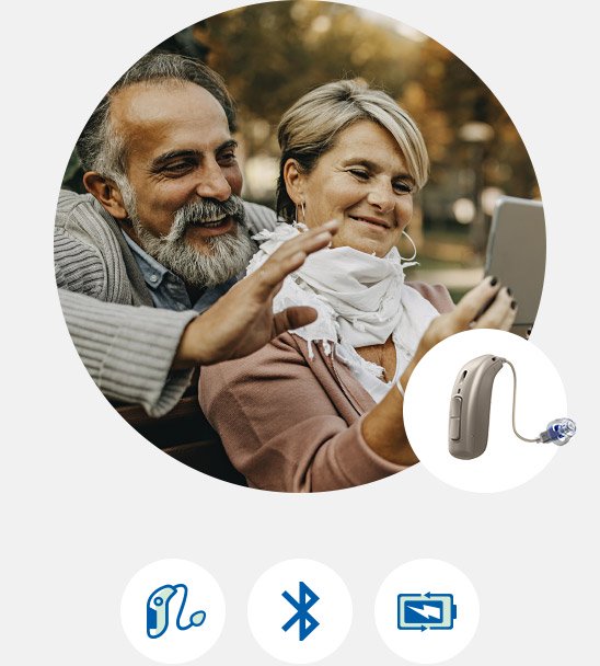 Smilende par sidder udendørs og ser på en tablet, med et høreapparat og ikoner for funktioner som Bluetooth og streaming i forgrunden.