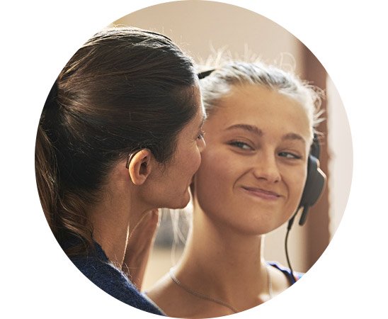 Eine Frau trägt ein diskretes Hörgerät hinter dem Ohr, während sie mit einer anderen Person spricht, die Kopfhörer trägt; die Szene deutet auf einen Hörtest in einer Hörklinik hin.