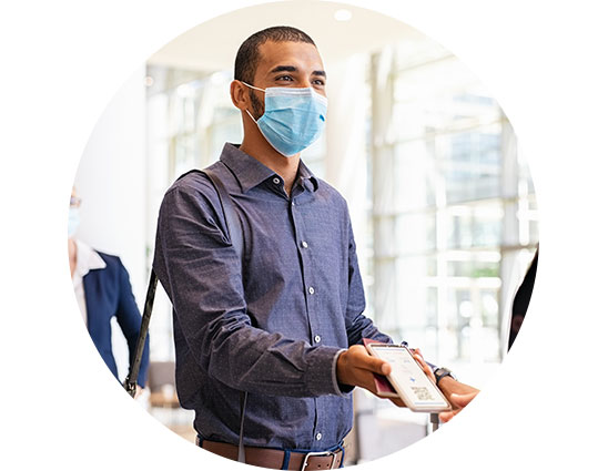 A person wearing a button-up shirt hands over a device, possibly a smartphone, to another individual in a modern, well-lit environment with large windows and visible individuals in the background.