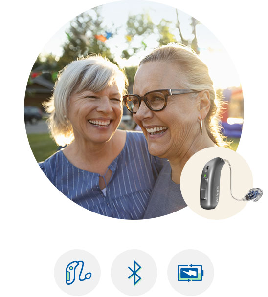 To høreapparater fra Oticon vises i forgrunden, ledsaget af ikoner for Bluetooth, streaming og lydindstillinger. I baggrunden ses to kvinder udendørs med sollys i fokus.