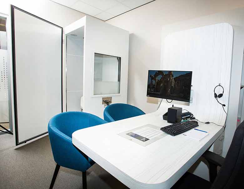 Een moderne audiologiepraktijk met een witte tafel, twee blauwe stoelen, een computer met hoorset, en een nabijgelegen geluidscabine voor gehoortests in een klinische inrichting.