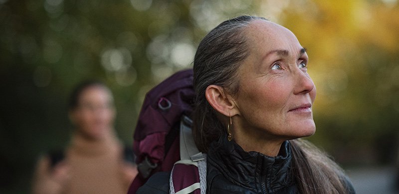 Eine Frau mit langem Haar trägt eine schwarze Jacke und einen Rucksack, während sie in einer herbstlichen, bewaldeten Umgebung steht. Ihr Ohr ist im Fokus, kein Hörgerät erkennbar.