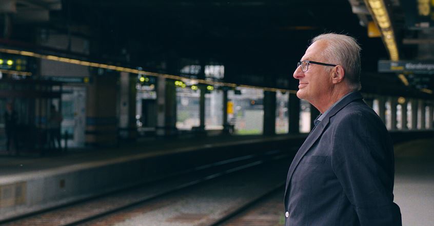 Ein älterer Mann in dunklem Anzug steht wartend auf einem Bahnsteig, umgeben von Schienen, beleuchteten Anzeigen und Bahnhofselementen unter einer überdachten Struktur. Kein Text oder erkennbares Hörgerät sichtbar.