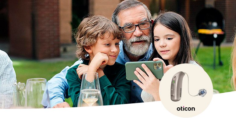 Eine Familie sitzt an einem Tisch mit Getränken im Garten und betrachtet gemeinsam ein Smartphone. Im Vordergrund ist ein Hörgerät von Oticon abgebildet, passend zum Thema Hörversorgung.