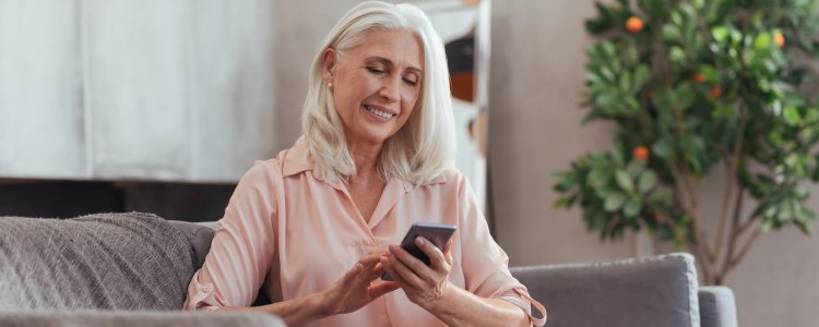 Lächelnde ältere Frau mit schulterlangem grauem Haar sitzt auf einem Sofa und blickt auf ihr Smartphone, im Hintergrund steht eine grüne Zimmerpflanze.