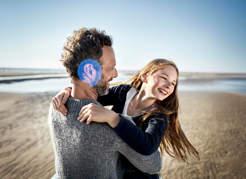 Grossvater trägt lachende Enkelin am Strand – Symbol für Nähe und gemeinsames Hören über Generationen hinweg