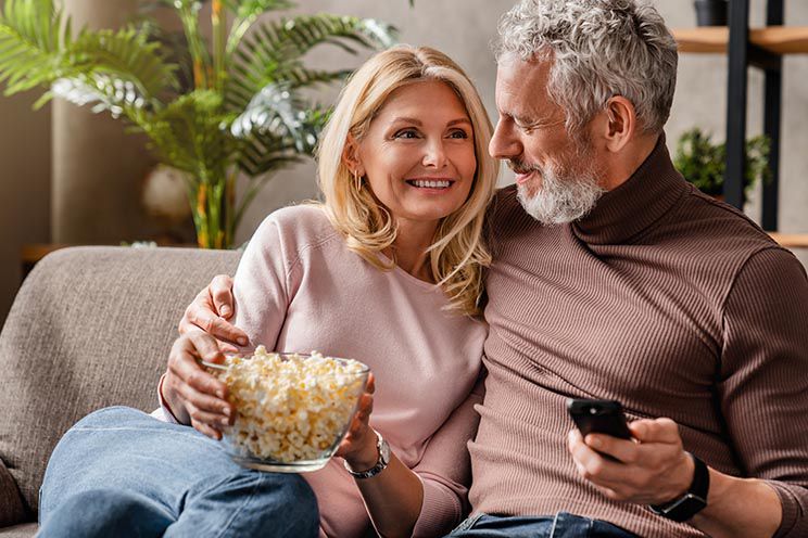 Ein älteres Paar sitzt auf einem Sofa; sie hält eine Schale Popcorn, während er mit einer Fernbedienung in der Hand sitzt. Im Hintergrund sind Pflanzen und Bücher zu sehen.