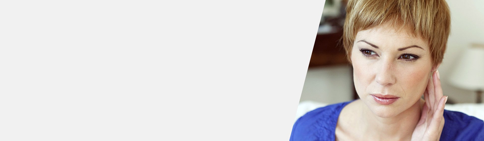 Eine Person (Gesicht unkenntlich gemacht) trägt ein blaues Oberteil, hebt die Hand zum Ohr. Im Hintergrund unscharfes Interieur mit Lampe. Kein sichtbares Hörgerät oder Text vorhanden.