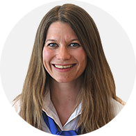 Eine Person mit mittellangem, glattem, braunem Haar trägt eine weisse Bluse mit einem blauen Schal. Das Gesicht ist unscharf. Der Hintergrund ist neutral und hell.