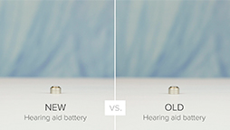 Eine neue Hörgerätebatterie (links) und eine alte Hörgerätebatterie (rechts) werden nebeneinander auf weissem Untergrund präsentiert. Text: 'NEW Hearing aid battery vs. OLD Hearing aid battery'. Hintergrund unscharf in Blautönen.