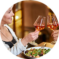 Zwei Personen stossen mit Weingläsern an, während sie an einem Tisch mit einem frischen Salat sitzen. Der Hintergrund zeigt ein warm beleuchtetes Restaurantambiente. Kein Text erkennbar.