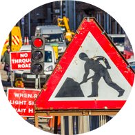 Baustellenschild mit arbeitender Figur vor urbaner Straßenszene mit Ampeln und Warnschildern. Symbol für Lärmbelastung und Bedeutung von Gehörschutz im Straßenverkehr.