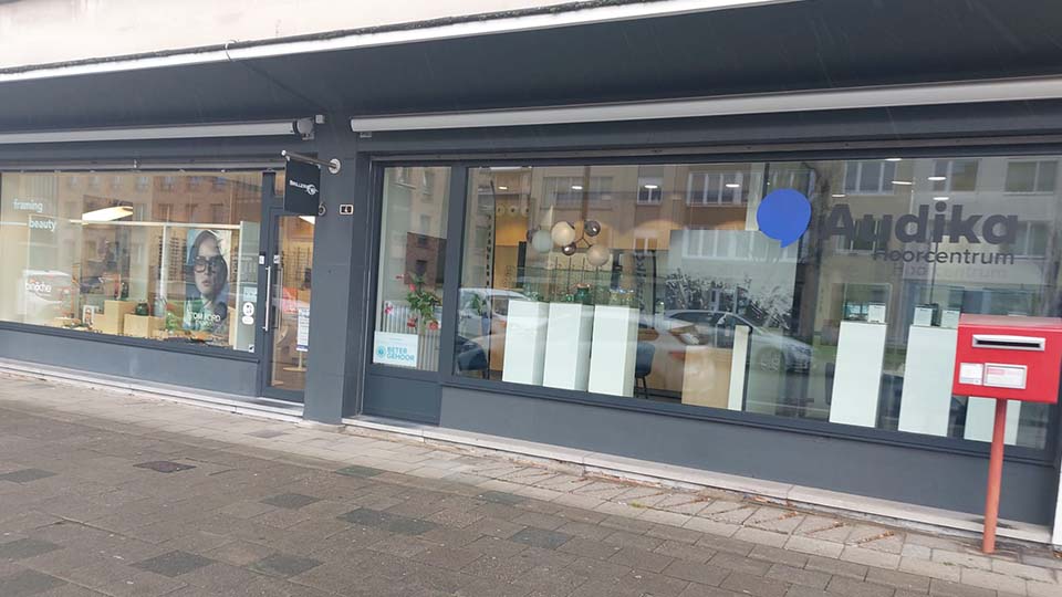 Façade d'une clinique Audika spécialisée dans des soins auditifs, avec de grandes vitrines affichant le logo bleu 'Audika Hoorcentrum'. Contexte urbain, boîte aux lettres rouge à droite.