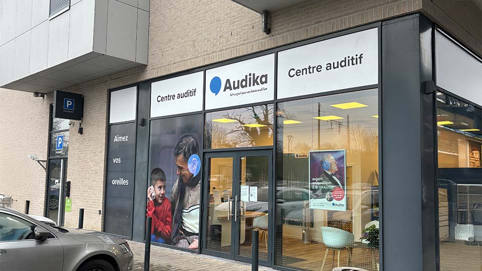 Façade d’un centre auditif Audika avec des vitrines affichant le texte 'Aimez vos oreilles'. Enseigne mentionnant 'Centre auditif'. Affiche promotionnelle à l'intérieur : 'expert en audition de qualité'. Un parking visible.