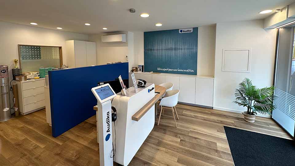 Un comptoir moderne d’accueil dans une clinique Audika, avec appareils électroniques, mobilier blanc et bleu, un sol en bois, une plante verte et le slogan « Venez opter pour une bonne audition ».