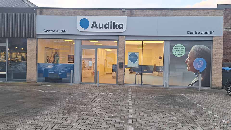 La façade d’un centre auditif Audika, avec vitres illuminées montrant l’intérieur moderne, porte 'Centre auditif' et slogan invitant au test auditif gratuit : 'Test auditif gratuit et sans engagement 0800 30 29 69 audika.be'.