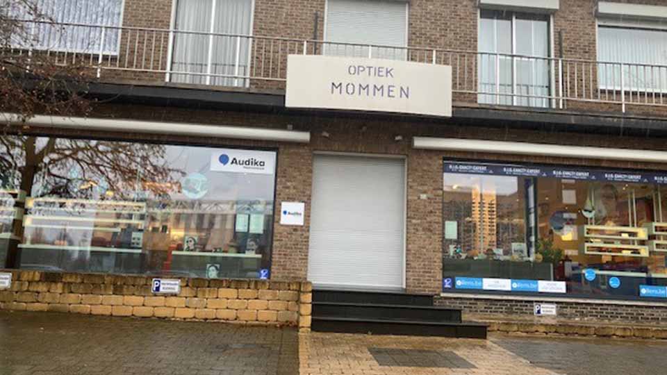 La façade d'une clinique Audika, spécialisée en soins auditifs, est située dans un bâtiment en briques. Enseigne 'Optiek Mommen', vitrines affichant le logo Audika et offres connexes.