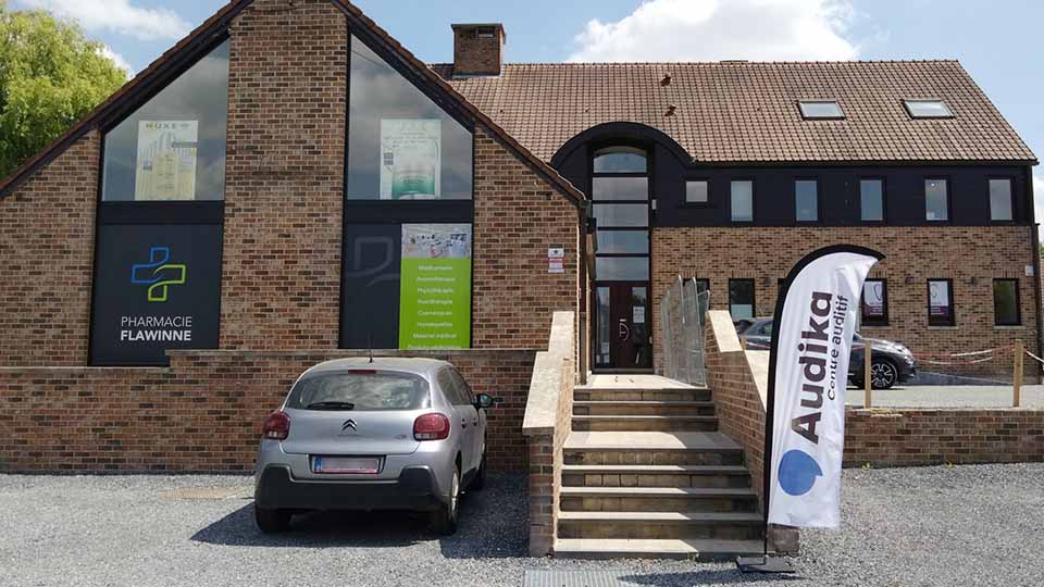 Un bâtiment en briques hébergeant une pharmacie ('Pharmacie Flawinne') et un centre auditif Audika. Une bannière Audika, une voiture grise, et un escalier dominent l'avant-plan.
