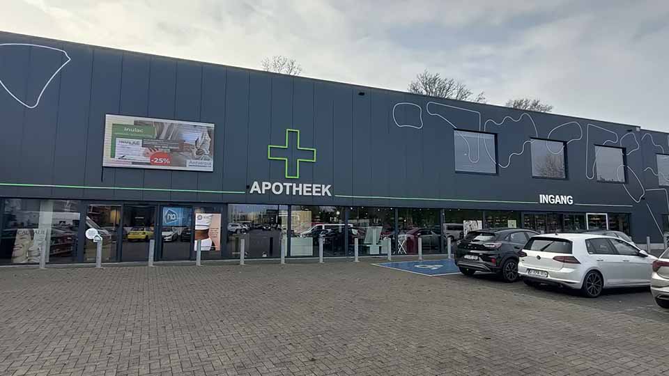 Façade d’une pharmacie moderne avec le mot ’APOTHEEK’ et une croix verte lumineuse. Plusieurs voitures stationnées devant l’entrée marquée ’INGANG’. Un panneau publicitaire indique ’-25% sur Nutricia.’