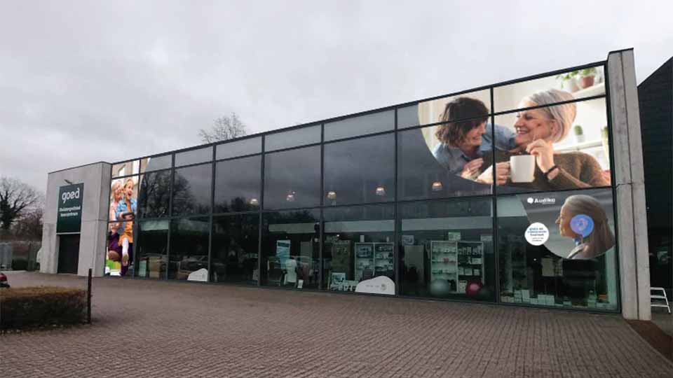 Un bâtiment moderne avec façade vitrée reflétant un ciel nuageux. La vitrine affiche des publicités d'Audika, une enseigne spécialisée en soins auditifs, et un panneau « goed Hoorcenters en Brillen ».