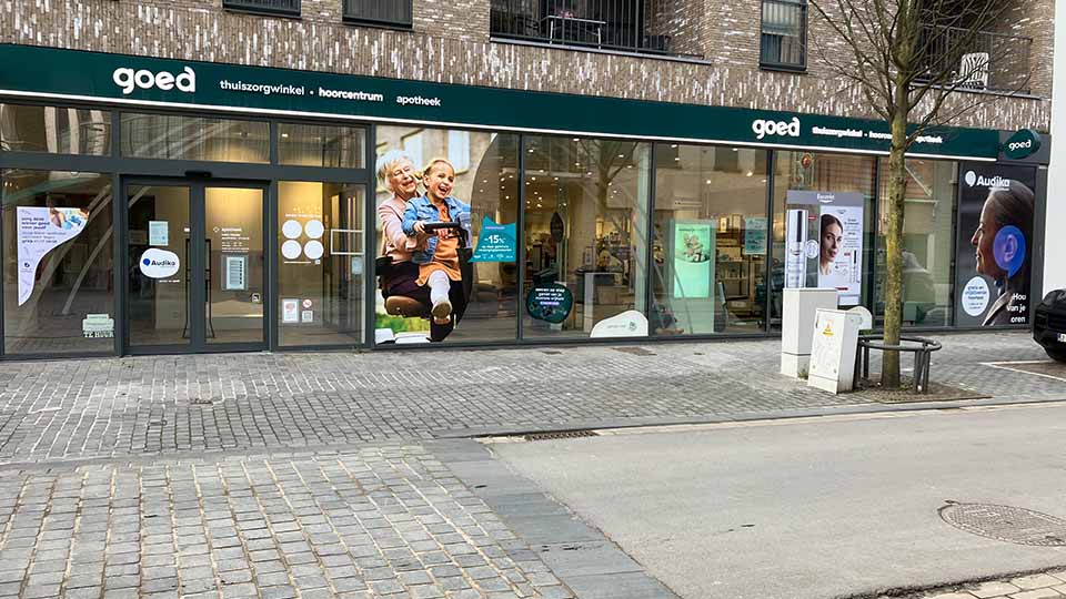 Façade d’un centre auditif et médical 'Goed' avec vitrine vitrée, affiches promotionnelles et logo Audika visible. Situé dans un quartier urbain moderne avec trottoir pavé.