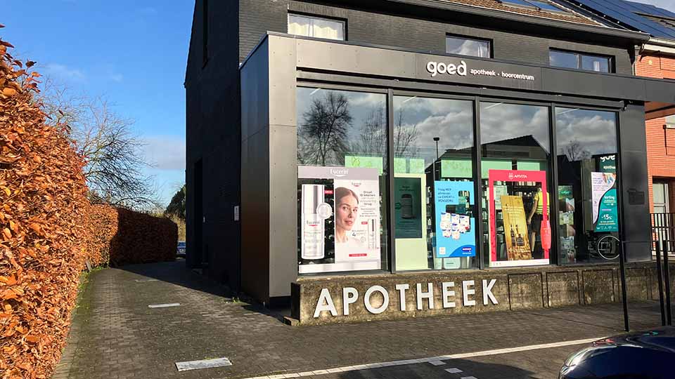Une façade moderne noire avec des vitrines affichant des publicités, identifiée comme 'goed apotheek - hoorcentrum'. Texte 'APOTHEEK' visible en bas. Environnement résidentiel avec haies et ciel dégagé.