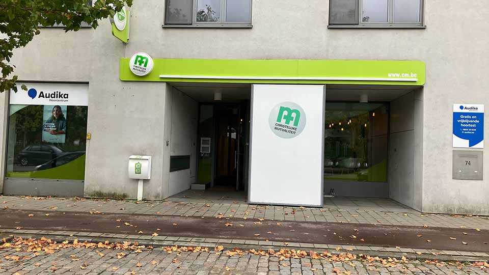 Une façade de bâtiment avec un centre Audika spécialisé en soins auditifs, affichant : « Hou van je oren » et un numéro d’appel (0800 30 523). Un arbre se tient devant.