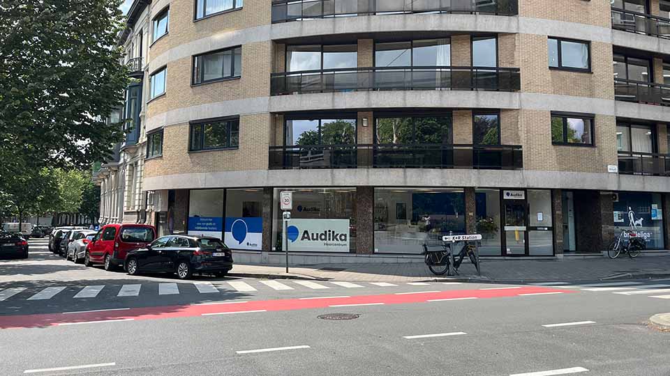 Une façade de clinique Audika spécialisée en soins auditifs, visible au rez-de-chaussée d’un immeuble moderne en briques jaunes avec vitrines, entourée de voitures garées et d’un passage piéton.